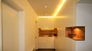 A narrow hallway with a wooden floor, white walls, and a built-in kitchen area with cabinets and a shelf. - Vacation apartment SeeFugium (Tegernsee)