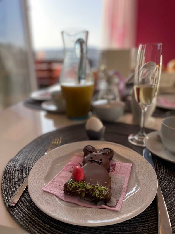 A dessert plate with a chocolate bunny and pistachio-topped pastry, a glass of white wine, and a pitcher of orange juice on a table with a woven placemat.