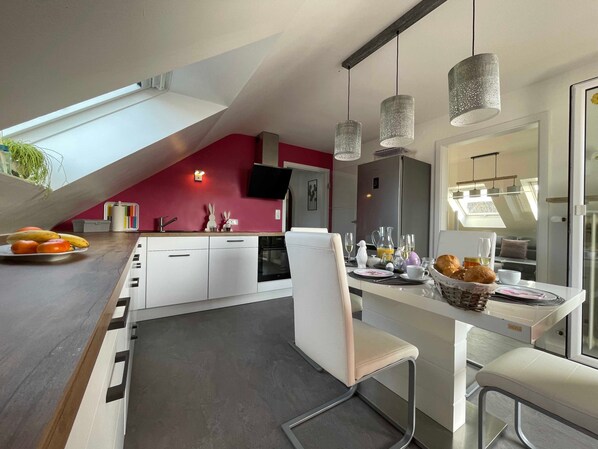 A modern kitchen with a dining area, a white chair, a table set for breakfast, and a large window.