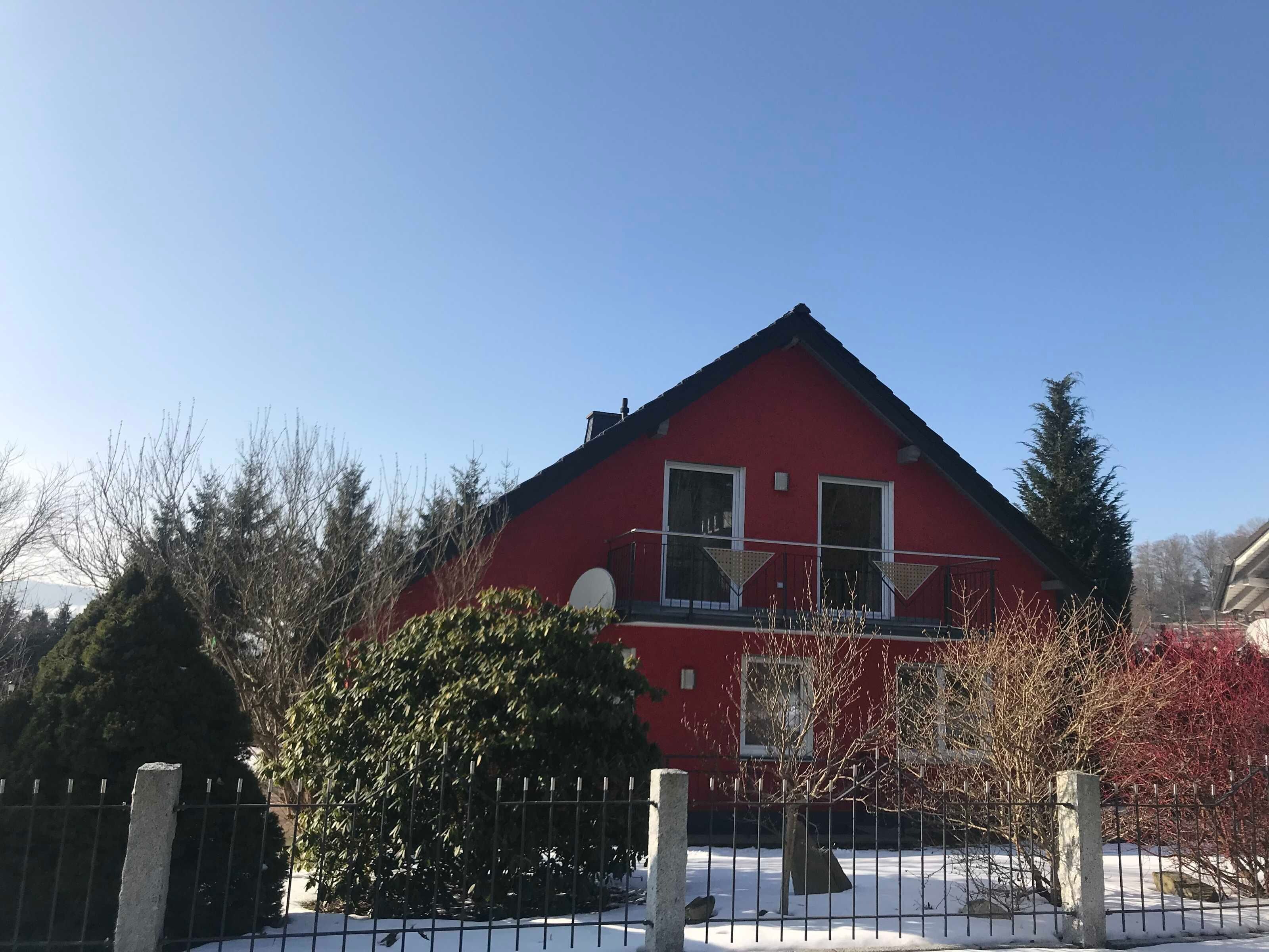 Una casa roja con techo triangular, rodeada por una valla metálica y suelo cubierto de nieve.