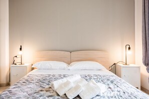 A neatly made bed with a patterned bedspread and white pillows, flanked by two bedside tables with lamps, in a simple room with light-colored walls.