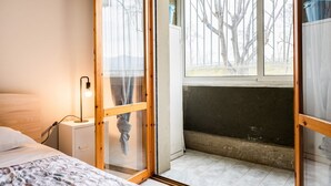 Ein Schlafzimmer mit Bett, Nachttisch, Lampe und Blick durch eine Glastür auf einen Außenbereich.
