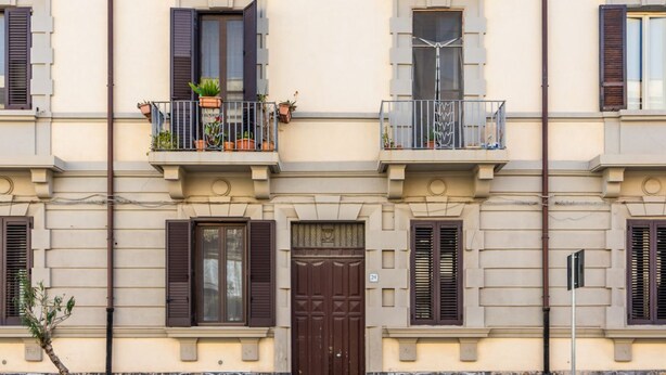 Un edificio con una puerta central marrón, varias ventanas con contraventanas y balcones con plantas en macetas.