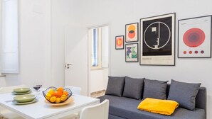 Een moderne woonkamer met een grijze bank, een witte tafel met een fruitschaal, en kunstwerken in lijsten aan de muur.
