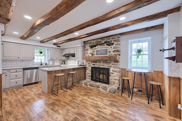 Una cocina con chimenea de piedra, vigas de madera, armarios blancos y una zona de bar.