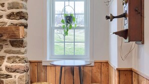 A room with a stone wall, a window with a view, a wooden table with two stools, and a wall-mounted light fixture. - Peaceful Hollow - Peaceful Hollow (Manchester)