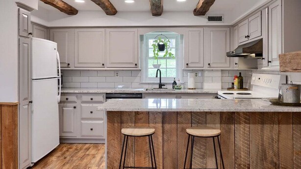 A kitchen with white cabinets, a wooden ceiling, a window with a plant, and a bar area with stools. - Peaceful Hollow - Peaceful Hollow (Manchester)