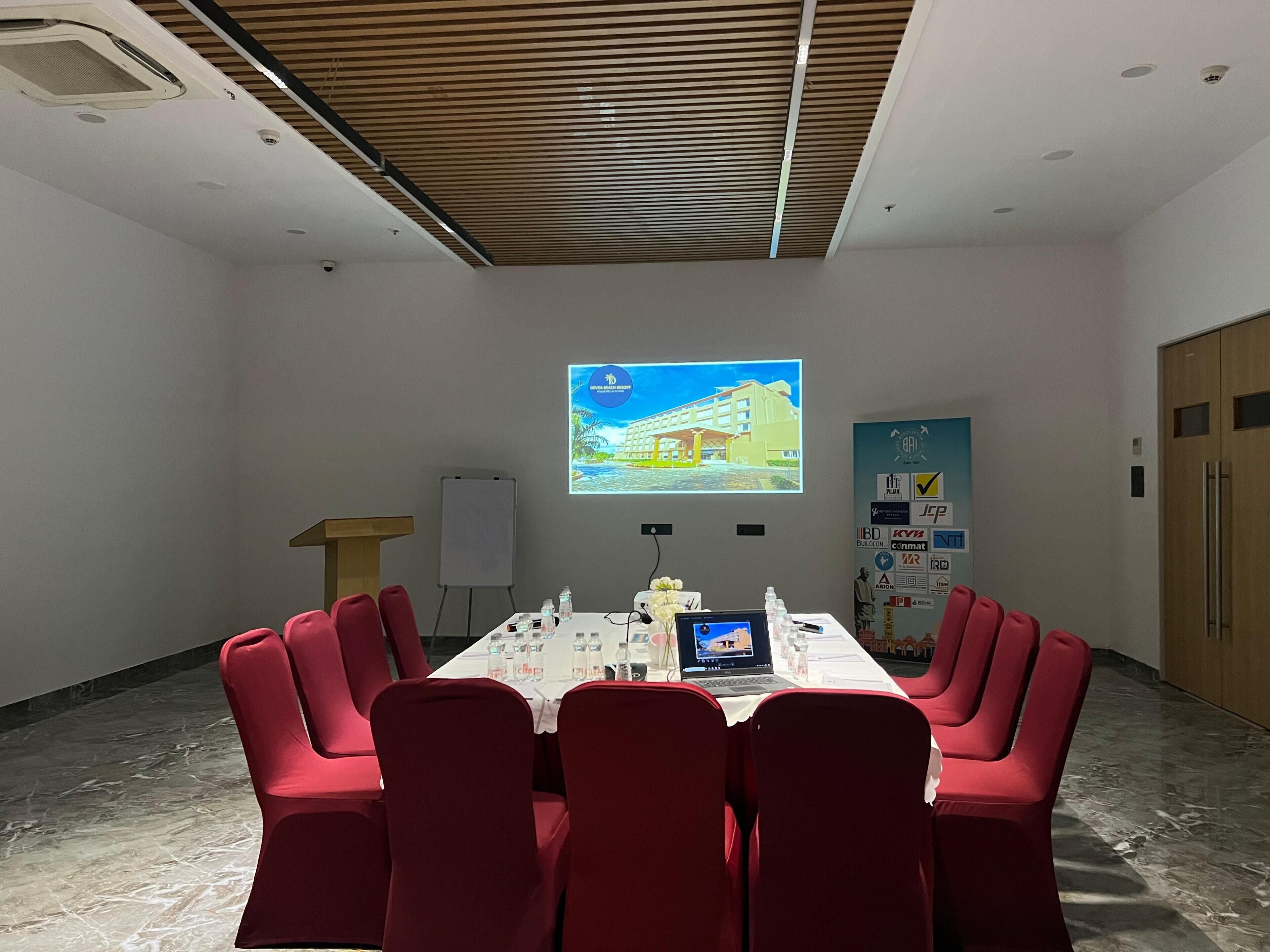 Un salón de conferencias con una larga mesa, sillas rojas, una pantalla de proyector y una pizarra.