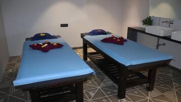 Two beds with pillows and decorative items, set against a geometric-patterned floor and a white wall, with a cabinet and sink nearby.