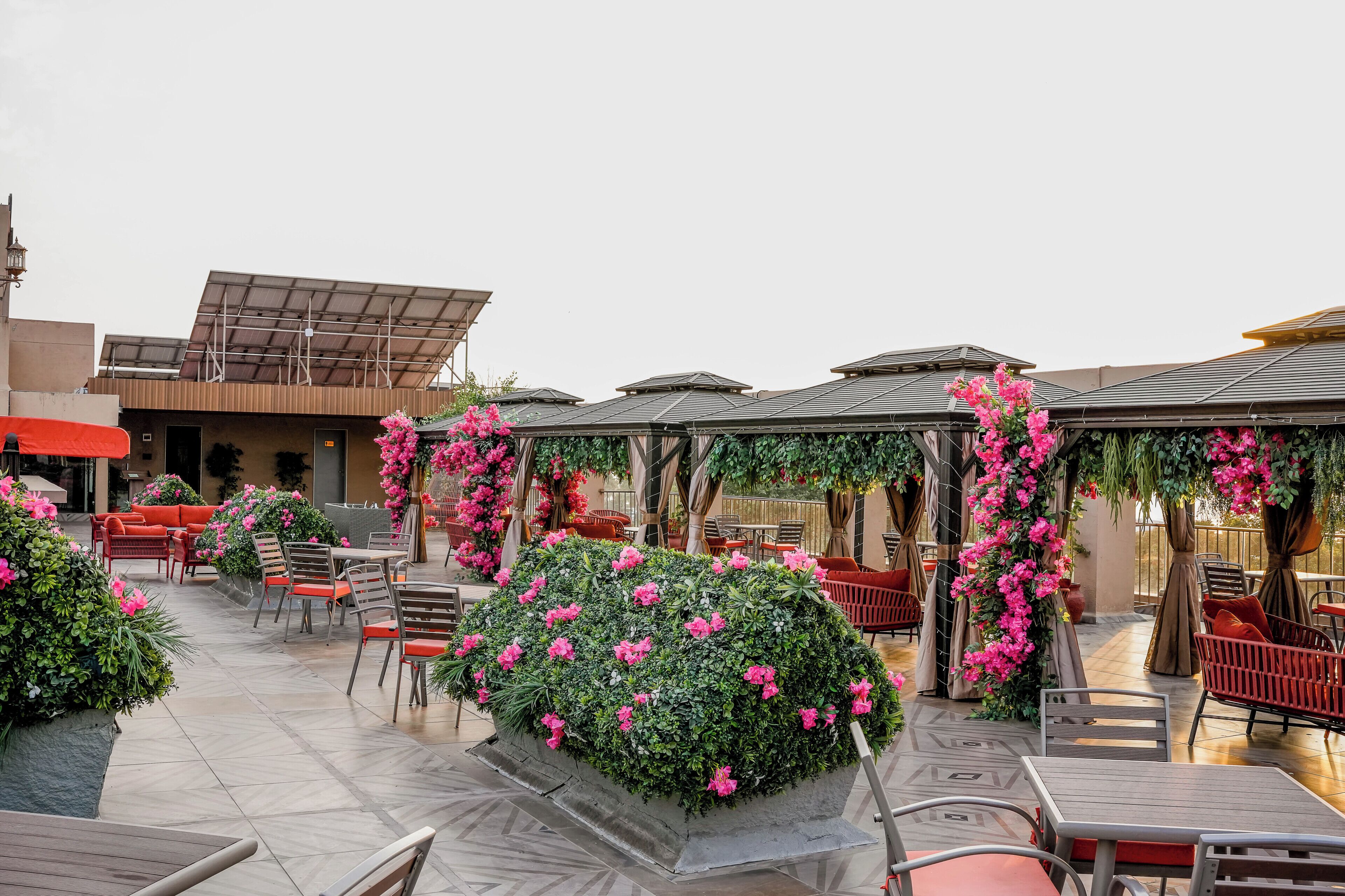 An outdoor seating area with tables and chairs, surrounded by greenery and pink flowers.