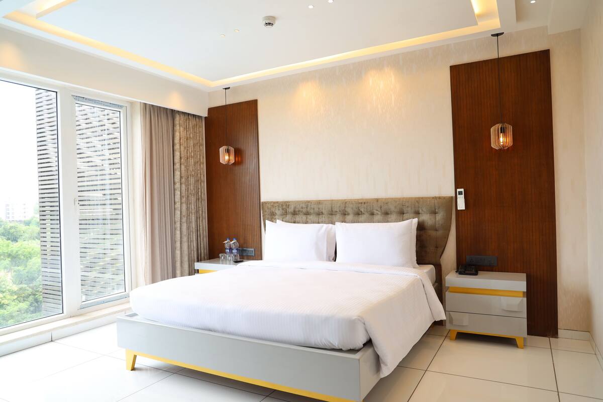 A modern hotel room with a large bed, a bedside table, a nightstand, and a view through floor-to-ceiling windows.