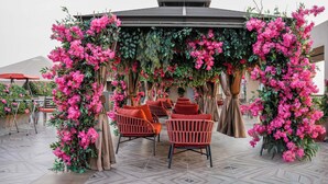 Et overdækket udendørs siddeområde med stole og borde, omgivet af frodige, lyserøde blomster.