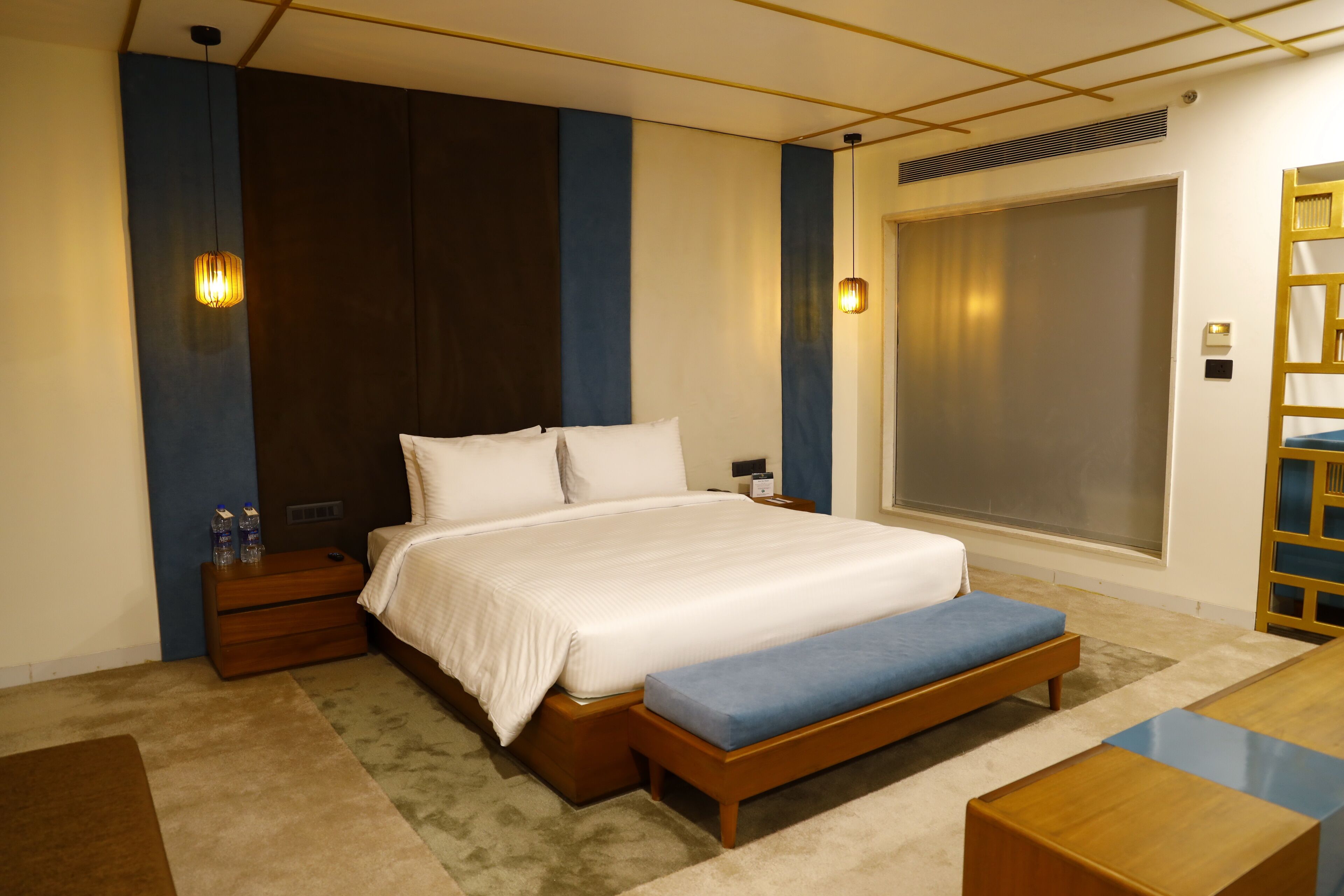 A modern hotel room with a large bed, bedside tables, a bench, and a view of the city.