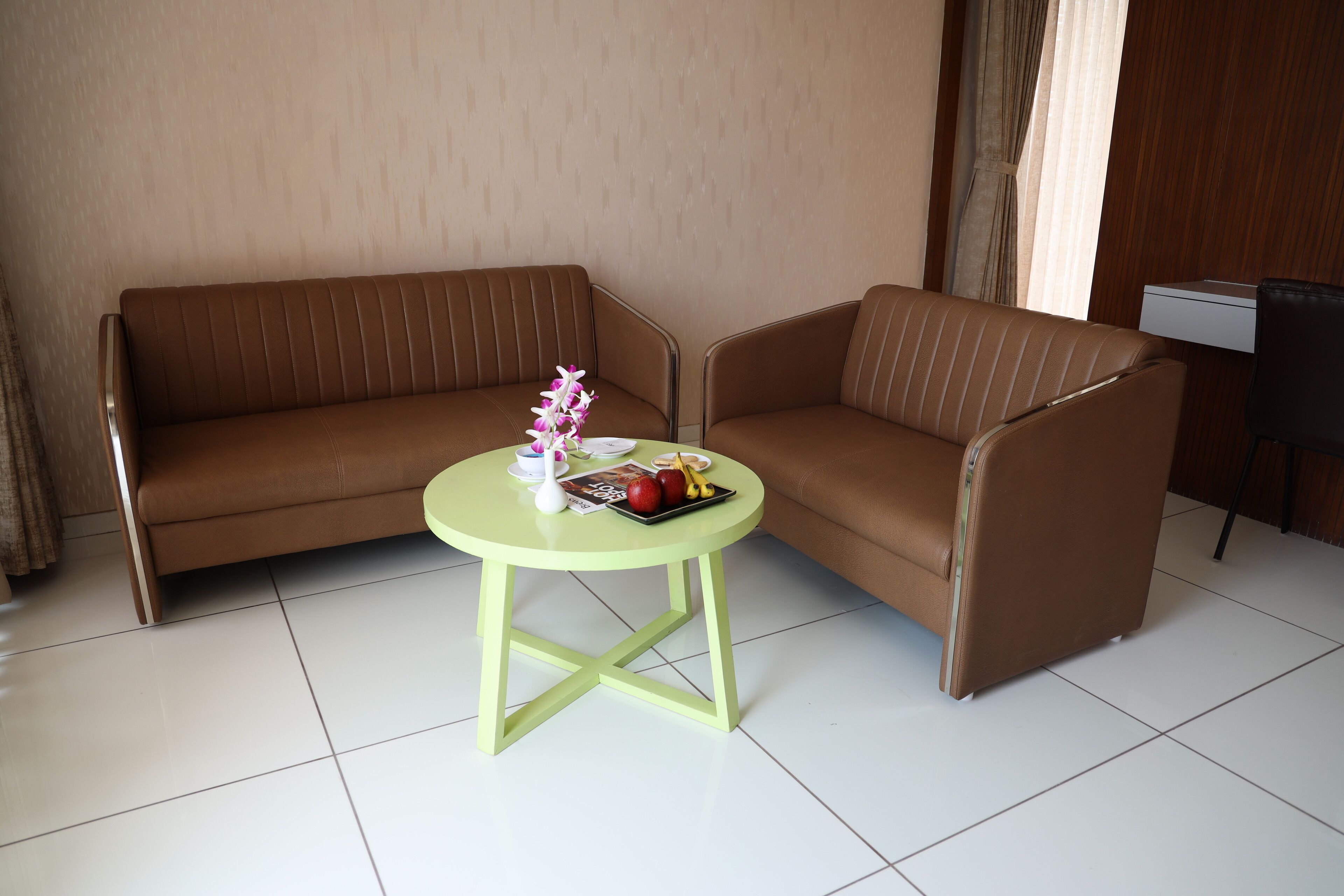 A room with a brown sofa and a small round table with a vase and fruit.