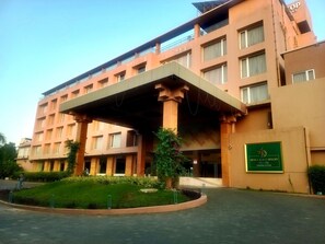 Un hotel de varios pisos con un acceso cubierto, un letrero verde y un área ajardinada con plantas.