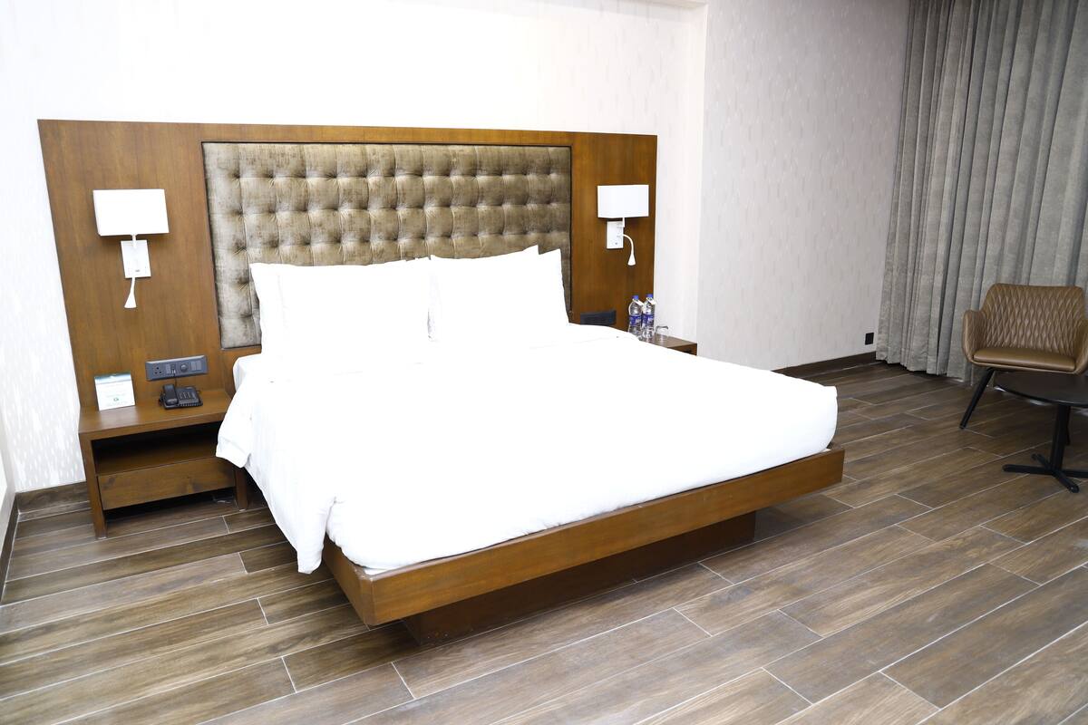 A hotel room with a large bed, wooden headboard, two bedside tables with lamps, a chair, and a curtain.