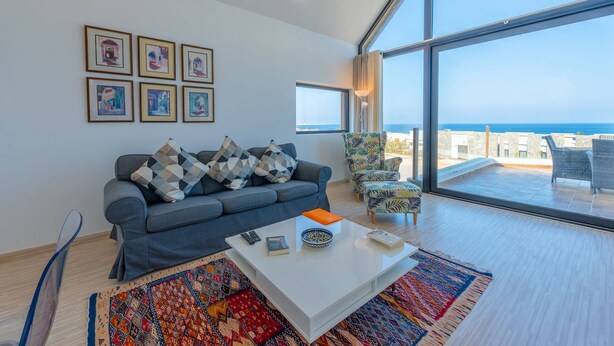 Una sala de estar moderna con un sofá gris, una mesa de centro y vistas al mar a través de amplios ventanales.
