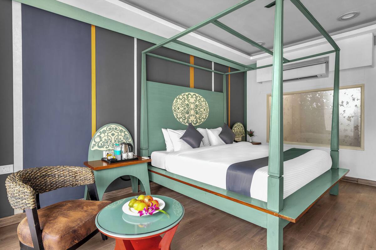 A four-poster bed with a green frame, a round table with a fruit bowl, and a chair with a woven backrest.