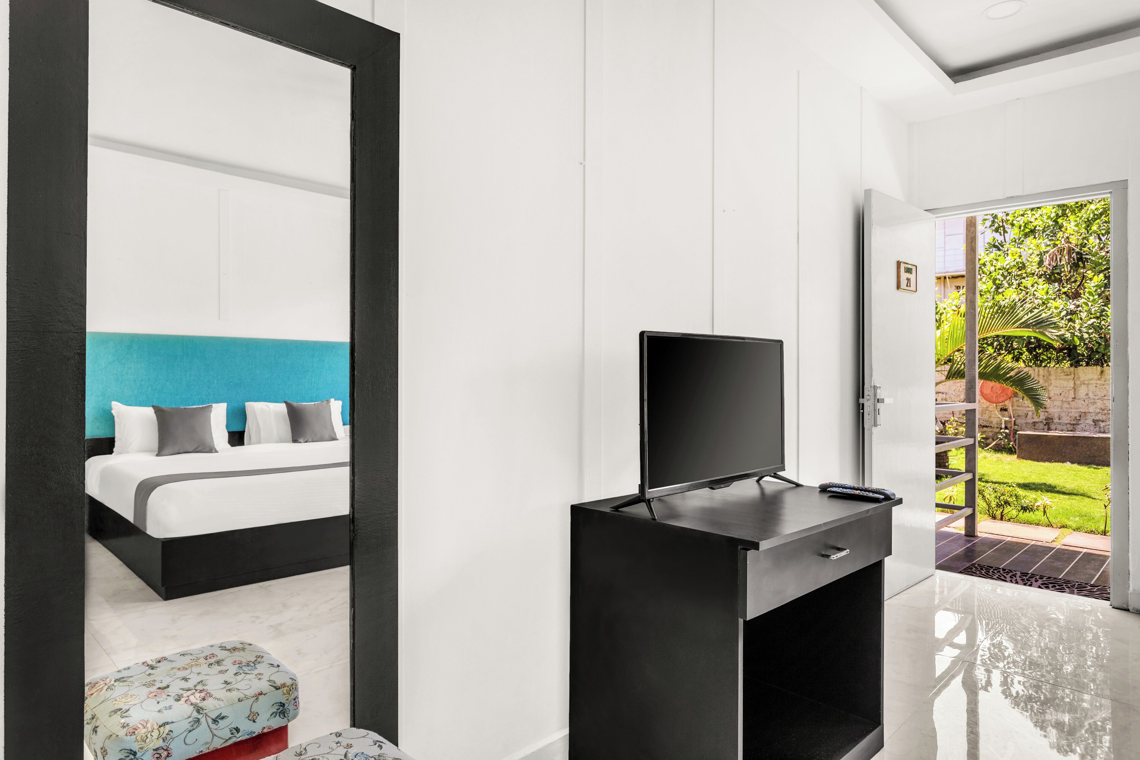 Una habitación de hotel moderna con una cama grande, un televisor sobre un pedestal negro y vistas a un jardín a través de una puerta abierta.