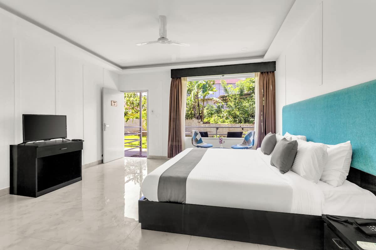 A modern hotel room with a large bed, a TV, and a balcony with a view of greenery.