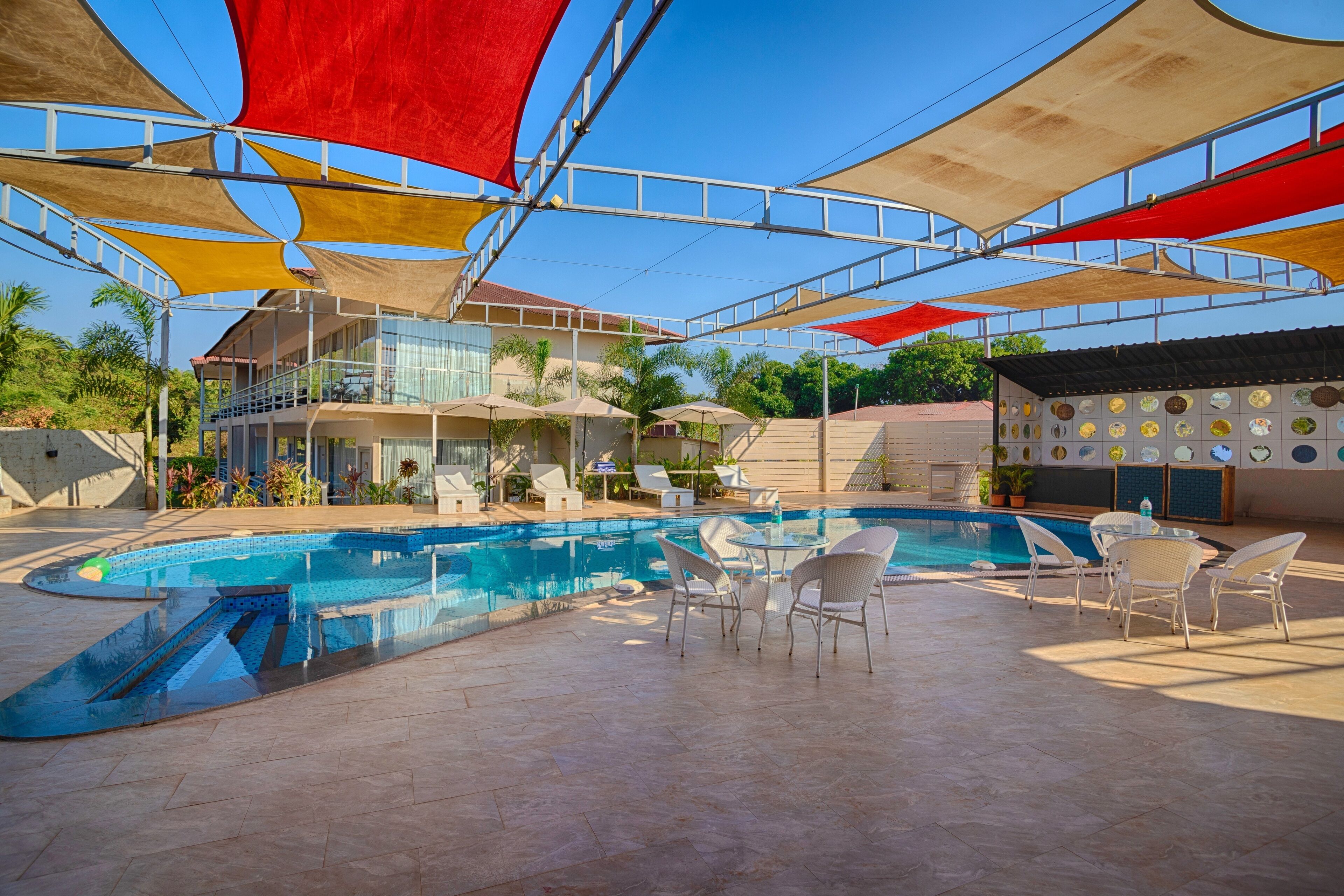A swimming pool with sunshades, white chairs, and a building with a balcony in the background.