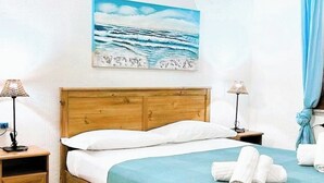 A hotel room with a wooden headboard, a bed with white bedding, a blue bedspread, two bedside lamps, a painting of the ocean, and a blue curtain.