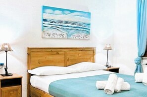 Ein Hotelzimmer mit einem hölzernen Kopfstück, einem Bett mit weißer Bettwäsche, einem blauen Überwurf, zwei Nachttischlampen, einem Bild des Ozeans und einem blauen Vorhang.