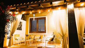 Un espacio exterior acogedor con una mesa y sillas, una ventana con contraventanas y una pérgola con luces colgantes.