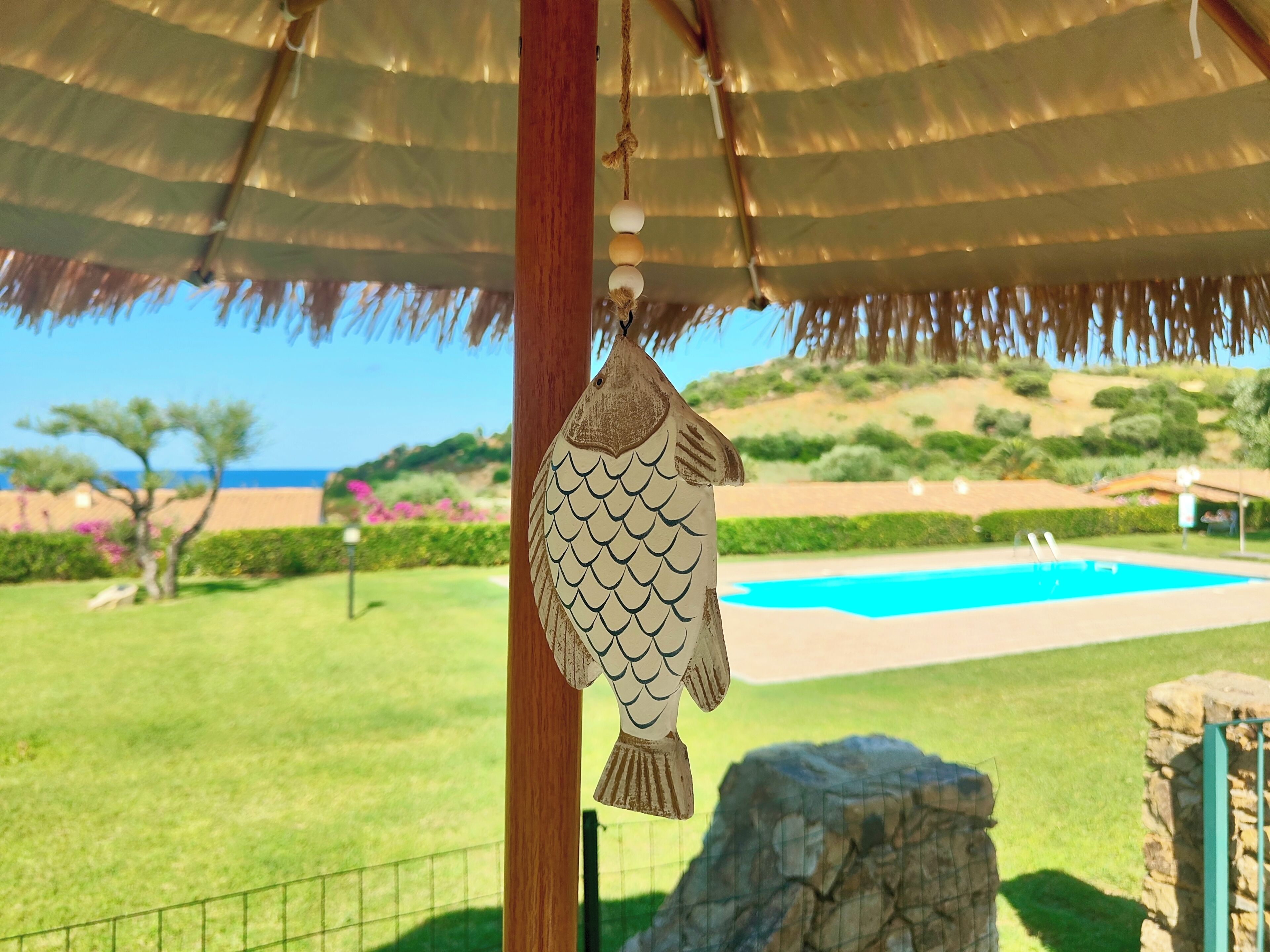 A fish-shaped decoration hanging from a structure with a view of a swimming pool and greenery.