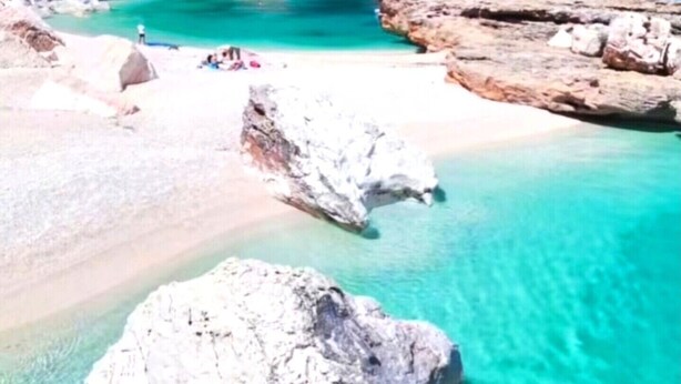 A coastal beach with clear turquoise water, large rocks, and a rocky cliff.