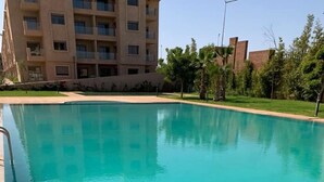 A clear swimming pool with a metal ladder on the left, surrounded by a well-maintained lawn and a multi-story building in the background. - La Perla de Marrakech (Marrakech)