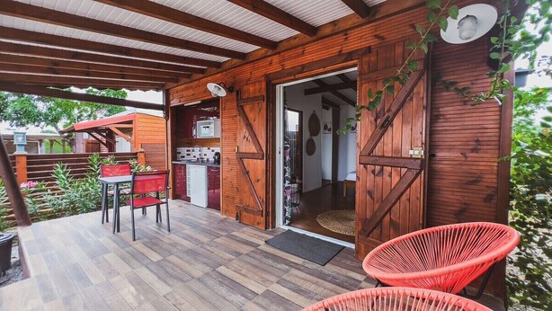 A wooden cabin with a porch, a dining area with a red table and chairs, and a kitchen visible through an open door. - Adrien Caraïbes • Domaine de Meilly (Saint-Louis)