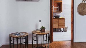 A room with a wooden floor, two round tables with metal wire frames, a flat-screen TV mounted on the wall, and a doorway leading to a kitchen area. - Adrien Caraïbes • Domaine de Meilly (Saint-Louis)