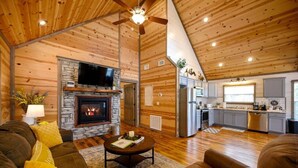 Un interior de cabaña acogedor con chimenea de piedra, televisor de pantalla plana, comedor y cocina.