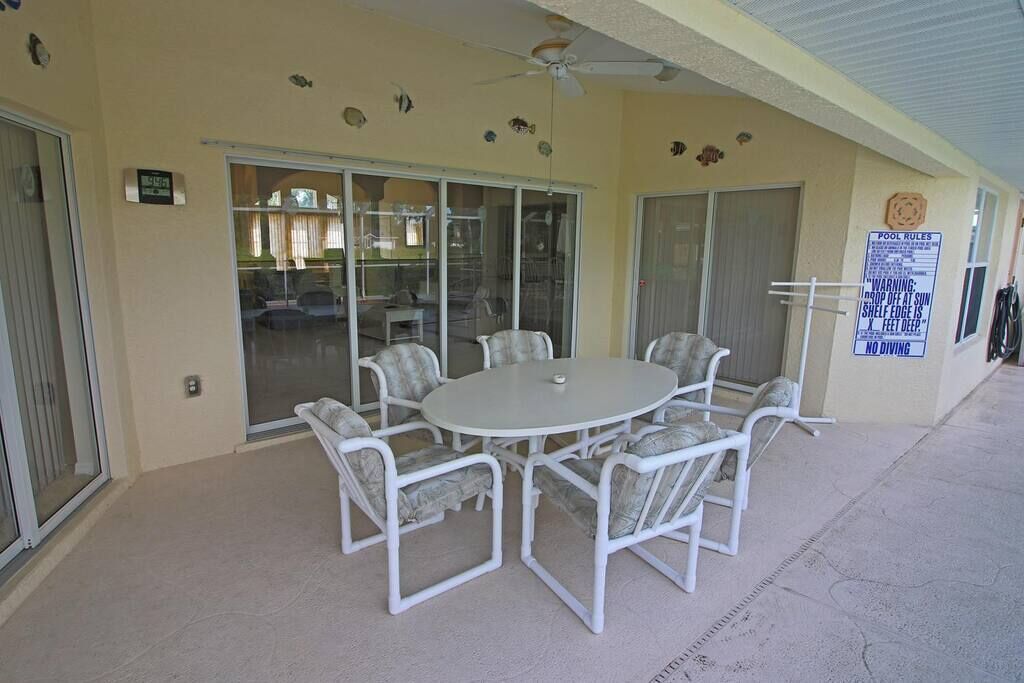 A covered outdoor seating area with a round table and chairs, a ceiling fan, and a pool rules sign.