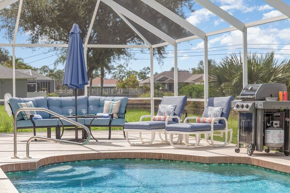 Un área junto a la piscina con una zona de estar techada, una parrilla y una piscina.