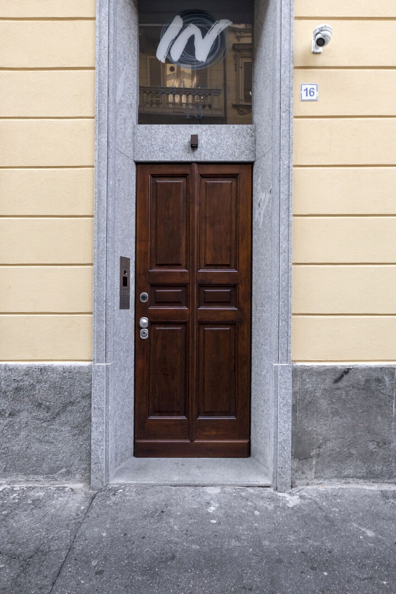 A double wooden door with a keypad lock, set in a stone frame, against a yellow wall with a number '16' sign.
