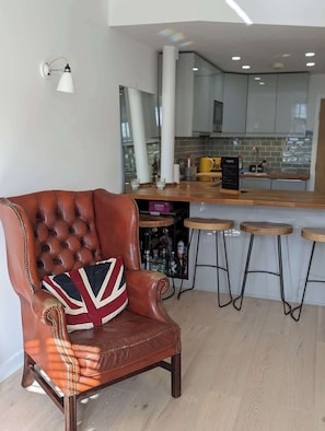 Un sillón de cuero con un cojín con la bandera del Reino Unido, una zona de bar con taburetes y una cocina moderna al fondo.