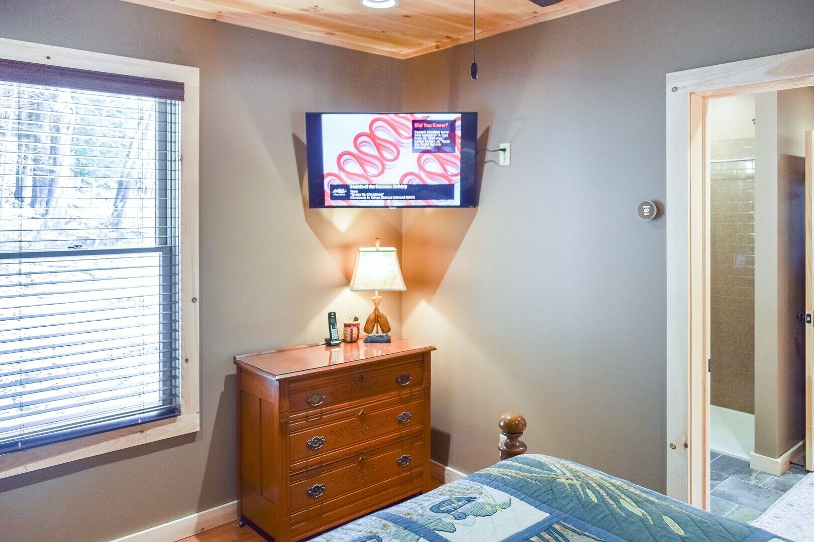 A bedroom with a wooden dresser, a bed with a blue quilt, a television mounted on the wall, and a window with blinds.
