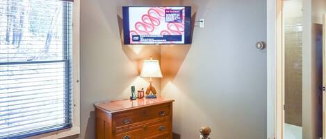 A bedroom with a wooden dresser, a bed with a blue quilt, a television mounted on the wall, and a window with blinds.
