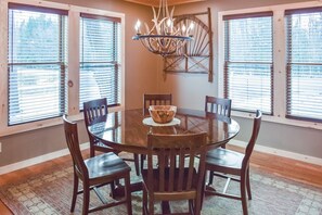 A dining area with a round table, six wooden chairs, a chandelier, and large windows with blinds. - Trails End House in Inlet (Inlet)