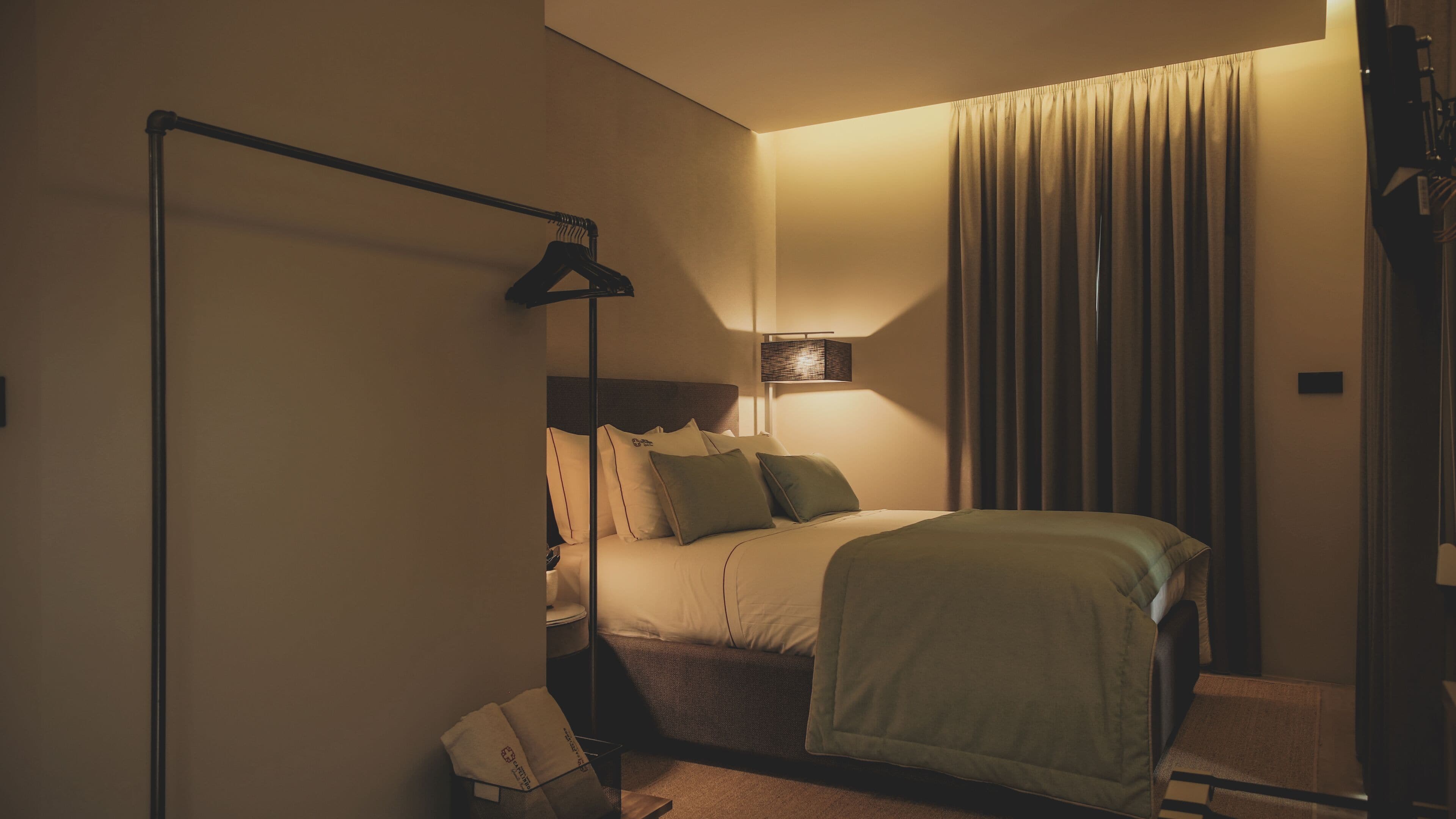 Quarto de hotel com cama, mesa de cabeceira, abajur e cortina.