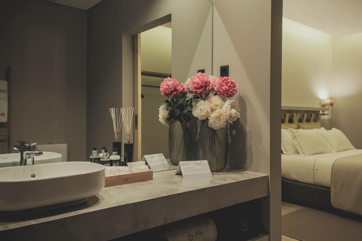 Casa de banho de hotel com lavatório, espelho e artigos de higiene. Há dois jarros grandes com flores e uma cama com cabeceira e almofadas.