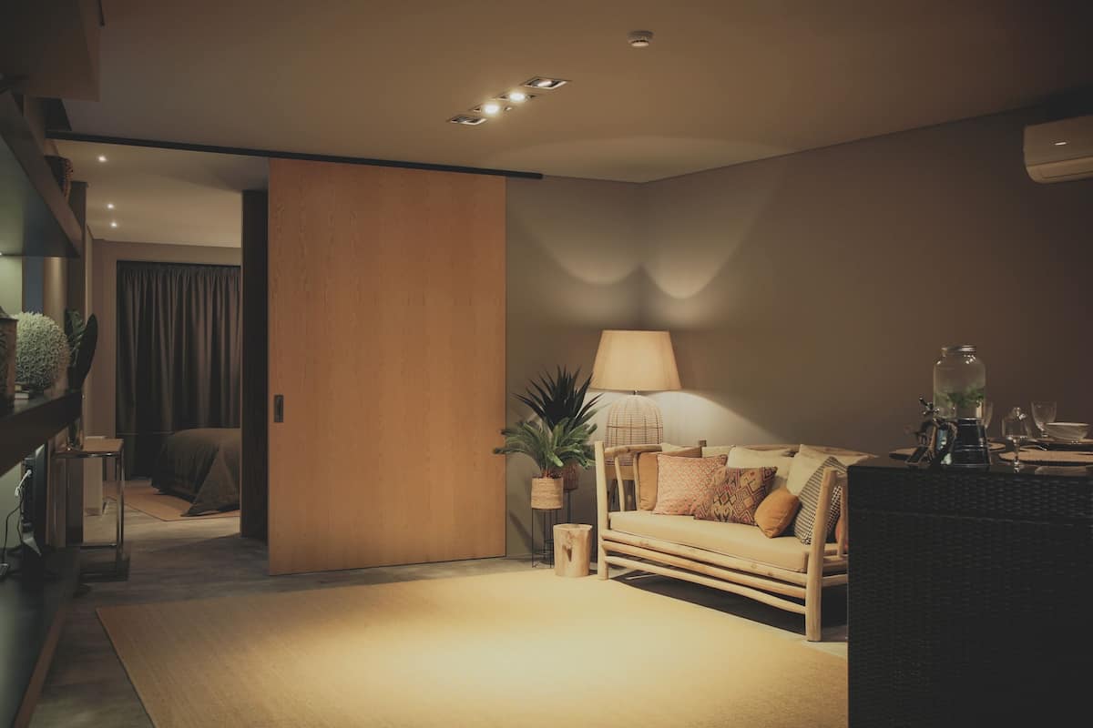 Sala moderna com chão de madeira, um sofá, uma mesa de centro e uma luminária. Há uma planta no chão e uma porta que dá para outro quarto.
