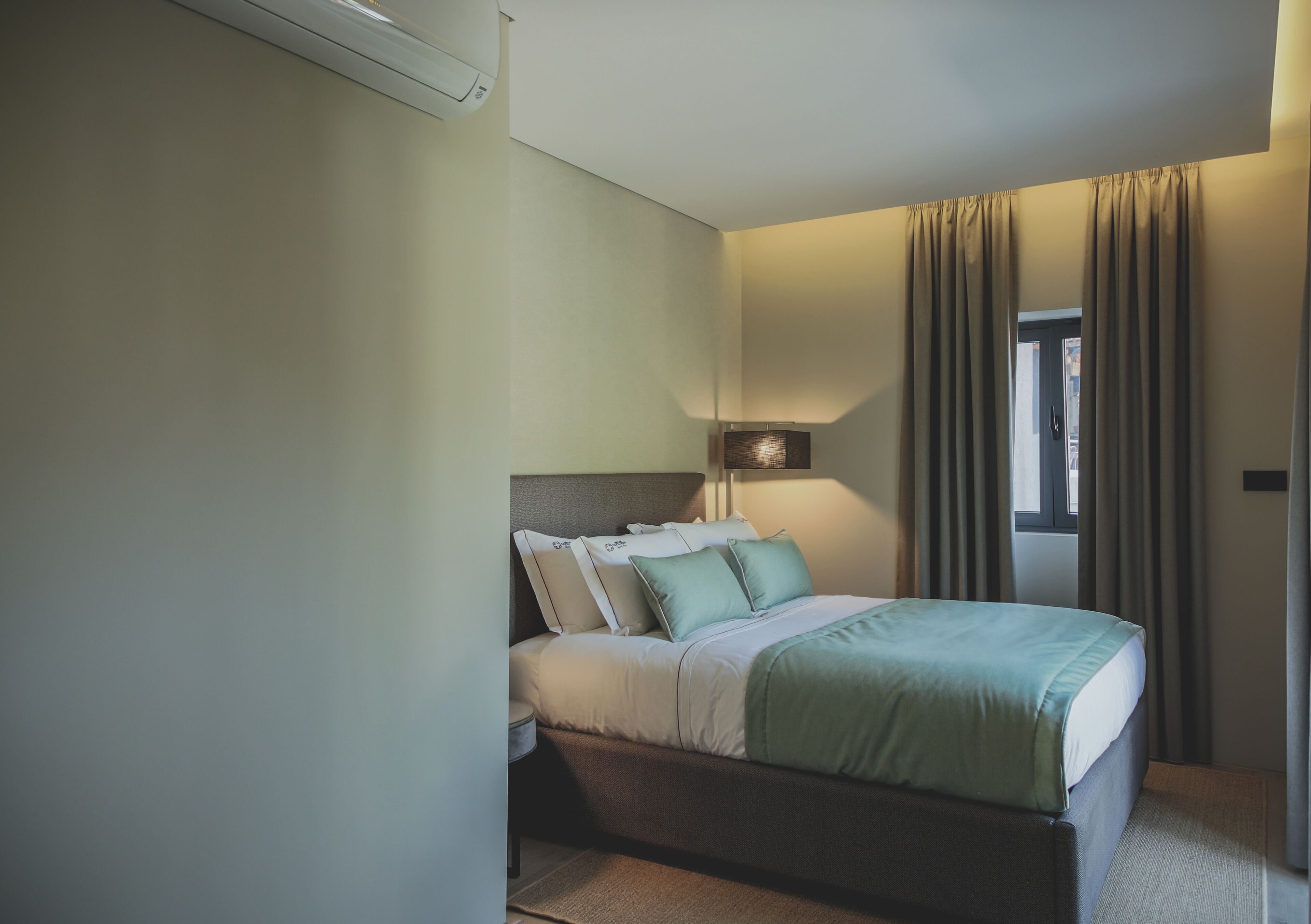 A bedroom with a bed, pillows, a bedside table, a lamp, and curtains.