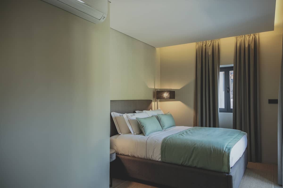 Quarto com cama, almofadas, mesa de cabeceira, abajur e cortinas.