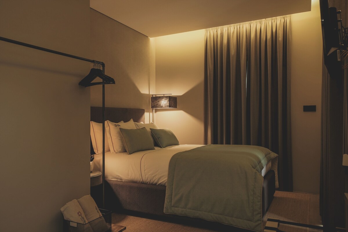 Una camera d'albergo con un letto, un comodino, una lampada, un lampadario e una finestra con tende.