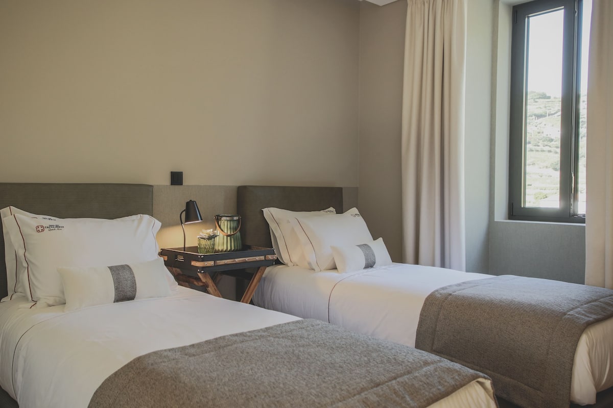 Quarto de hotel com duas camas, mesa de cabeceira com abajur e janela com cortinas.