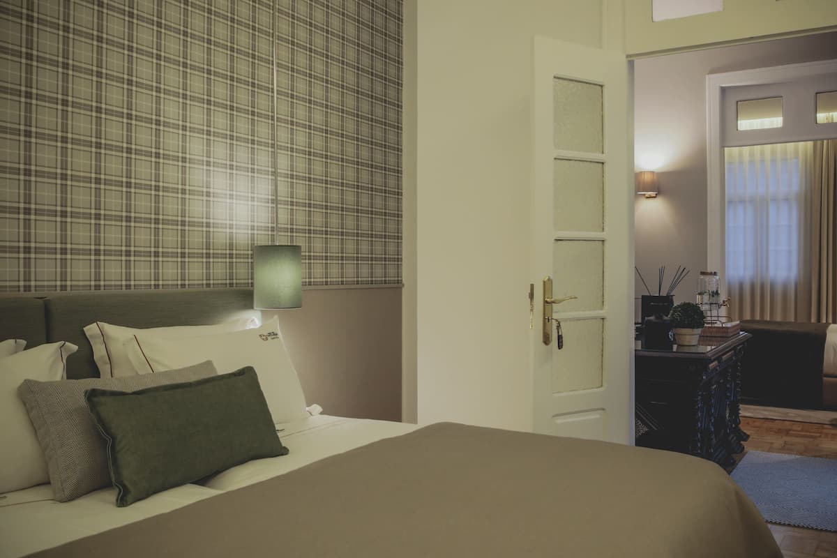 Quarto de hotel com cama, mesa de cabeceira, luminária, porta de acesso a um banheiro e vista para o banheiro através de uma janela.
