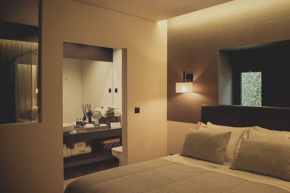 Quarto de hotel com cama, mesa de cabeceira, espelho e janela com vista para vegetação.